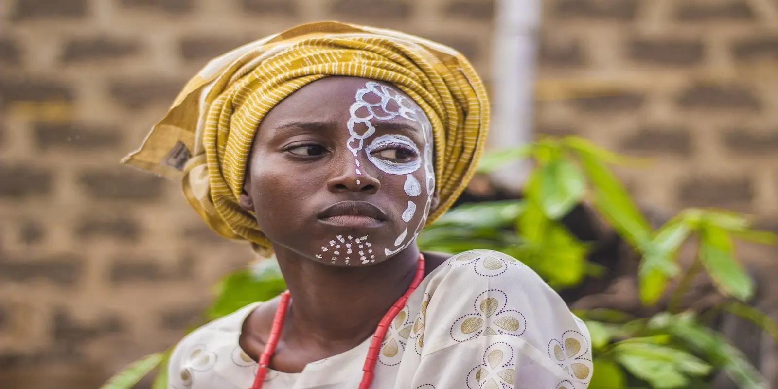 African woman with traditional white face paint wearing yellow headwrap and ceremonial attire against brick wall.