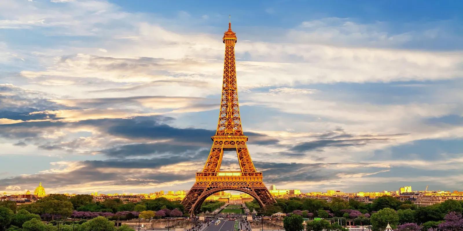 Eiffel Tower illuminated in golden light at sunset with Paris cityscape and dramatic clouds in background.