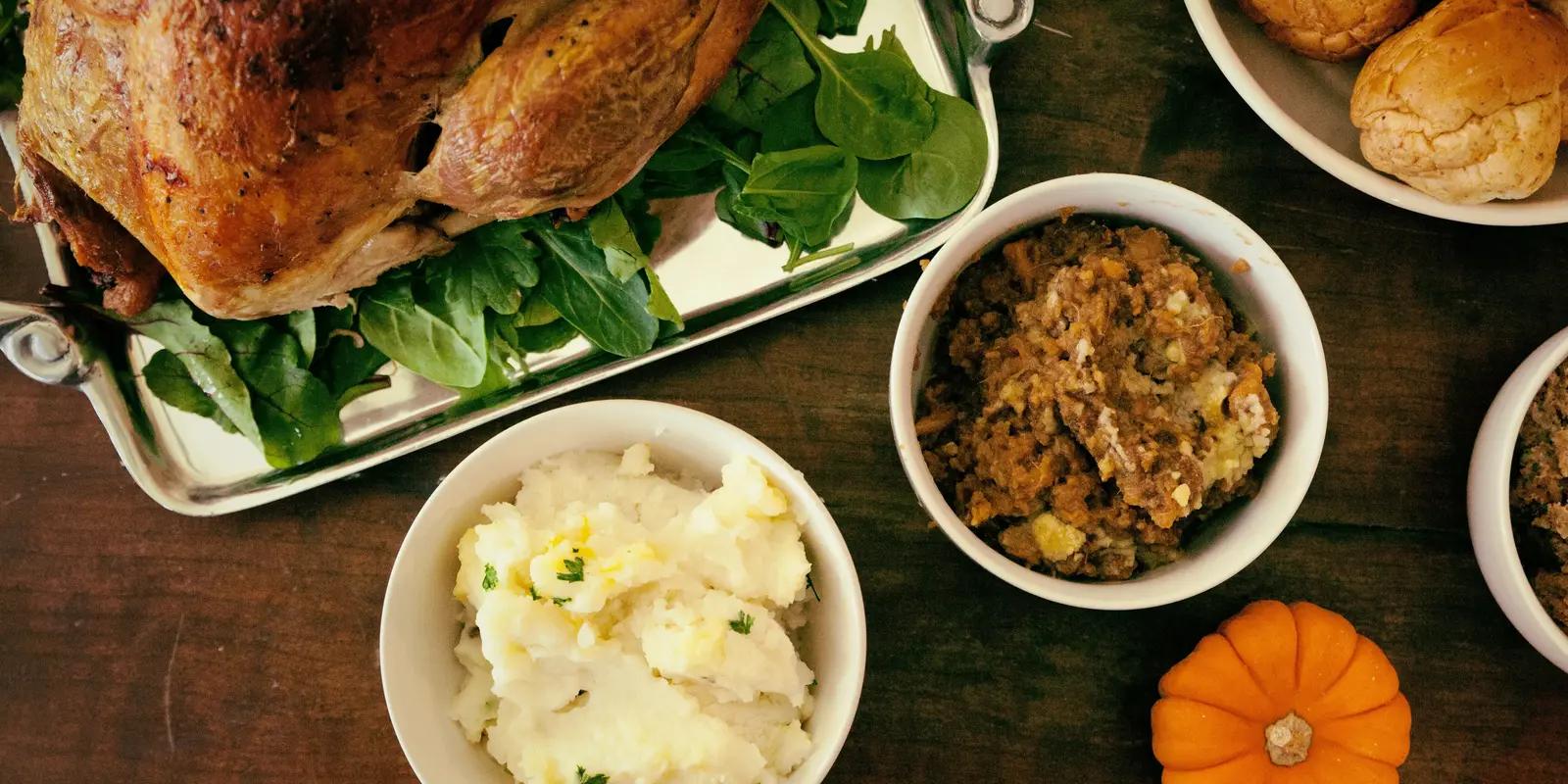 Thanksgiving dinner spread on wooden table featuring roasted turkey on platter with greens, mashed potatoes, stuffing in white bowls, and small decorative pumpkin.
