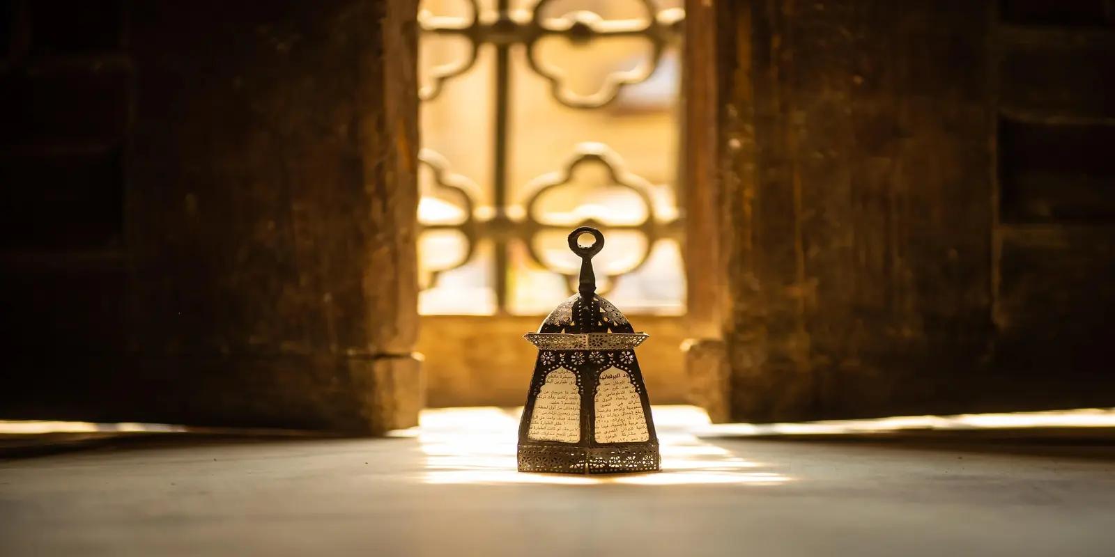 Ornate metal lantern with Arabic script illuminated in front of decorative window in historic mosque.