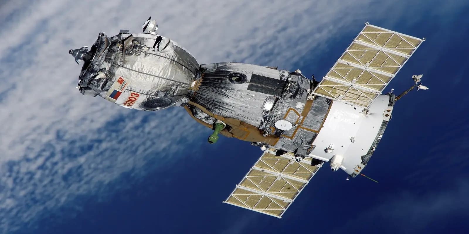 Russian Soyuz spacecraft in orbit above Earth with extended solar panels against space background.