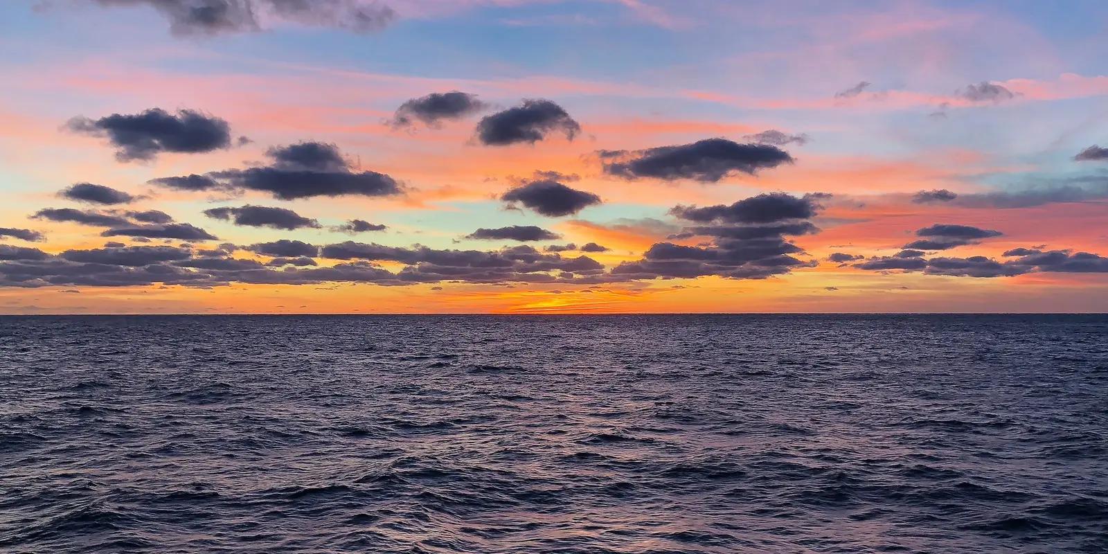 Ocean horizon at sunset with dark blue waters and colorful sky featuring orange, pink and purple clouds.