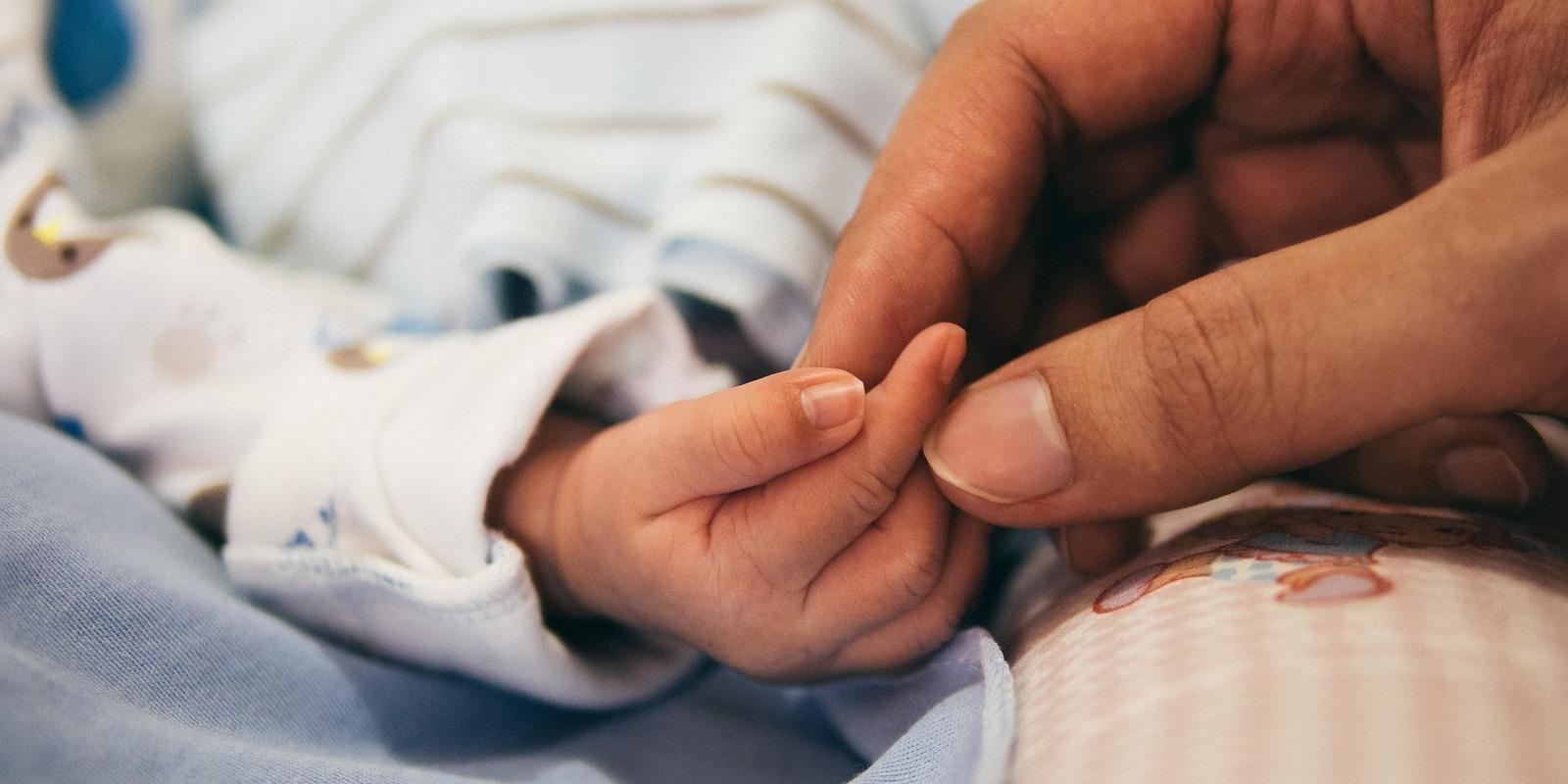 Adult hand gently holding tiny newborn baby's hand in hospital setting with blue and white clothing.