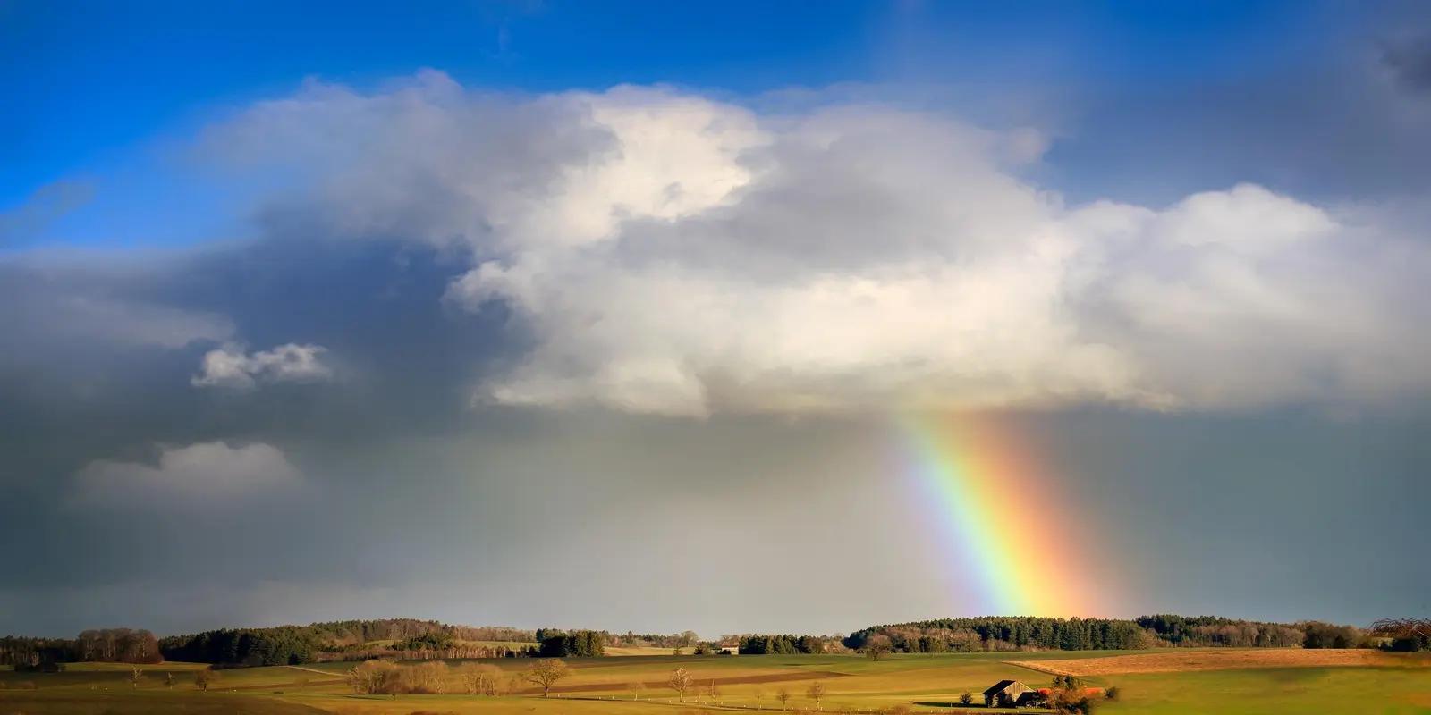 Vibrant rainbow stretching across dramatic cloudy sky over rural countryside landscape.