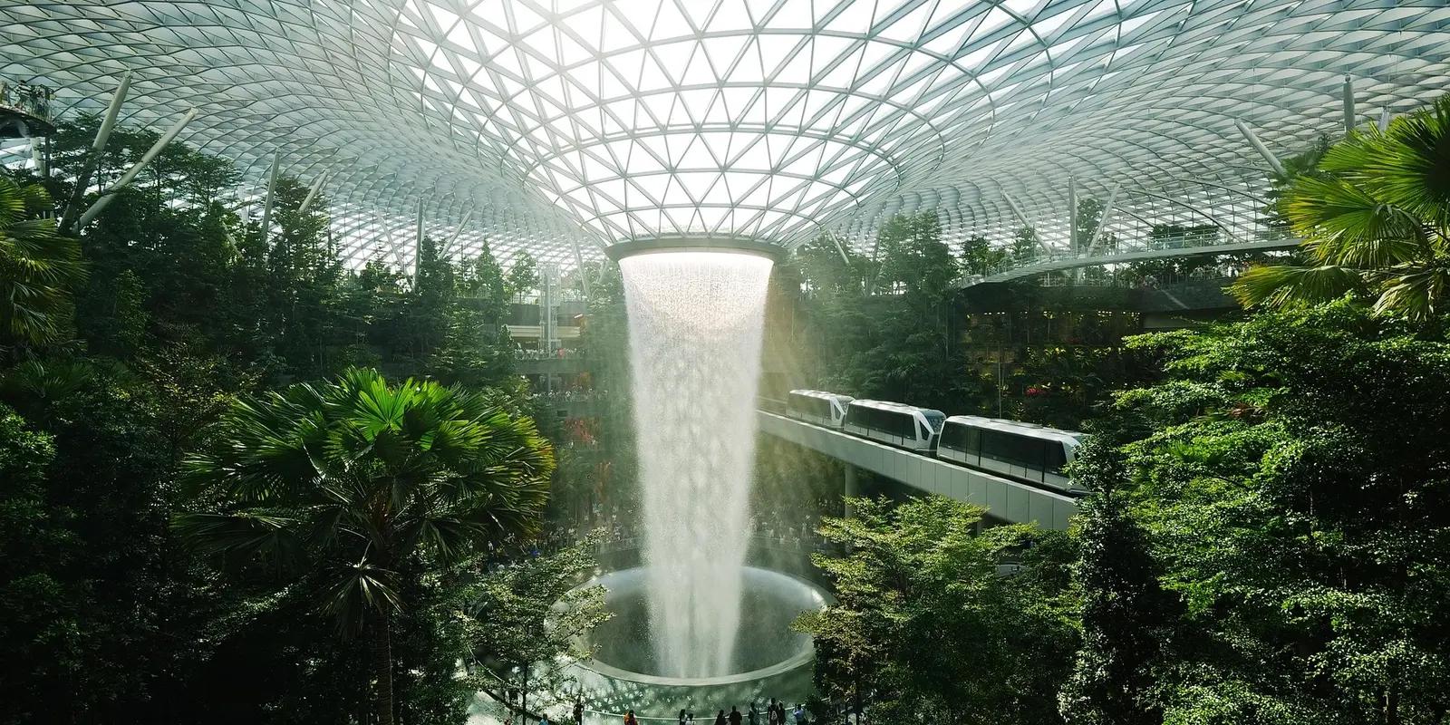 The HSBC Rain Vortex, the world's tallest indoor waterfall, cascades from a glass-domed ceiling into a circular basin at Jewel Changi Airport, surrounded by lush greenery and a sky train passing through.