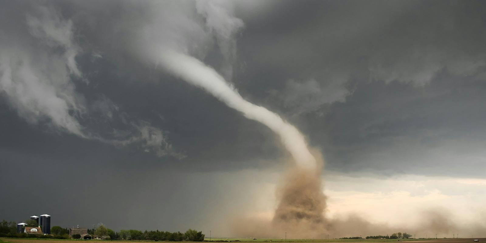 A powerful tornado spirals down from dark storm clouds over a rural landscape.