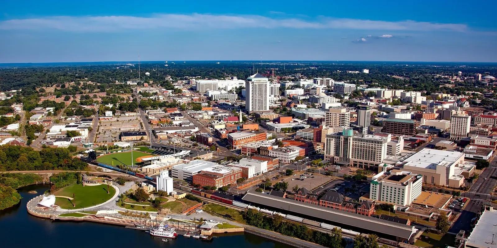Aerial view of downtown Montgomery, Alabama, showing the city skyline along the Alabama River with parks, stadiums, and historic buildings visible under a clear blue sky.