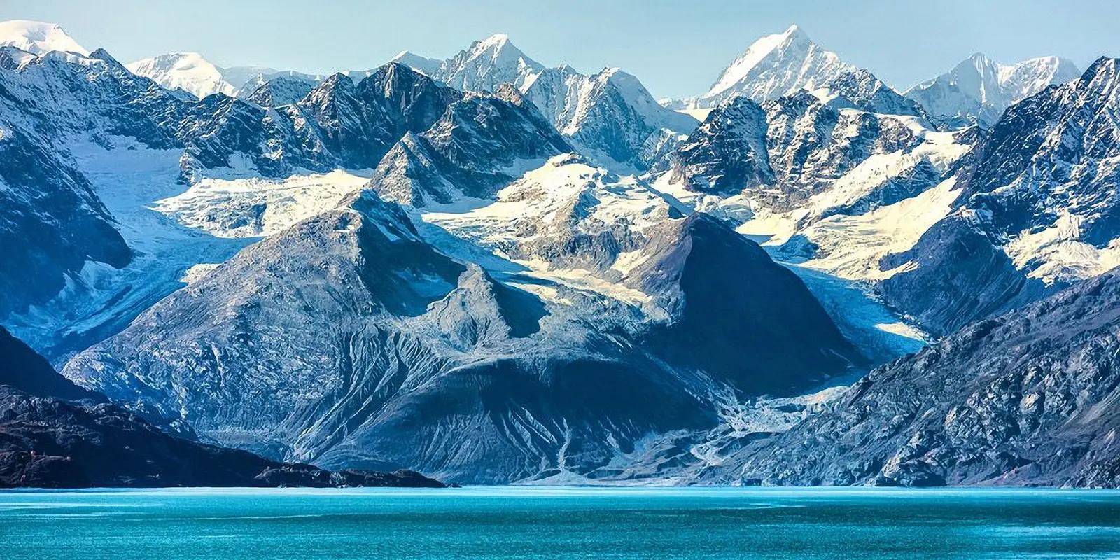 Dramatic Alaskan mountain range with snow-capped peaks and glaciers overlooking turquoise waters of Glacier Bay under clear blue skies.