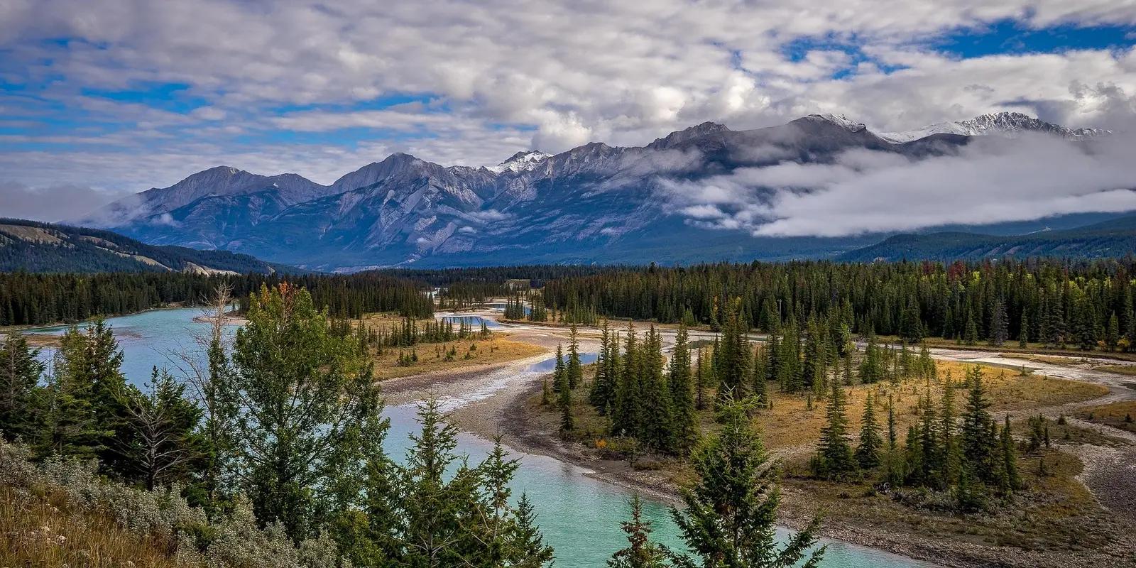 A breathtaking view of Jasper National Park, showcasing a turquoise river meandering through a forested valley, with towering mountains partially covered in mist and clouds in the background.