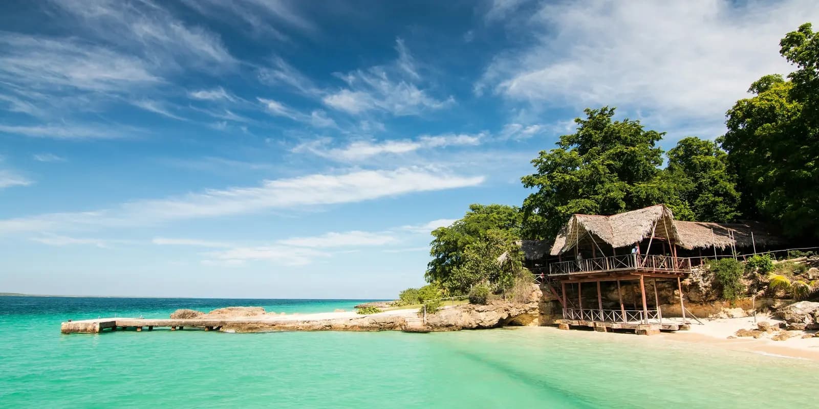 Tropical beach in Cuba with crystal-clear turquoise waters, a wooden pier, and rustic thatched-roof huts surrounded by lush green trees under a bright blue sky.