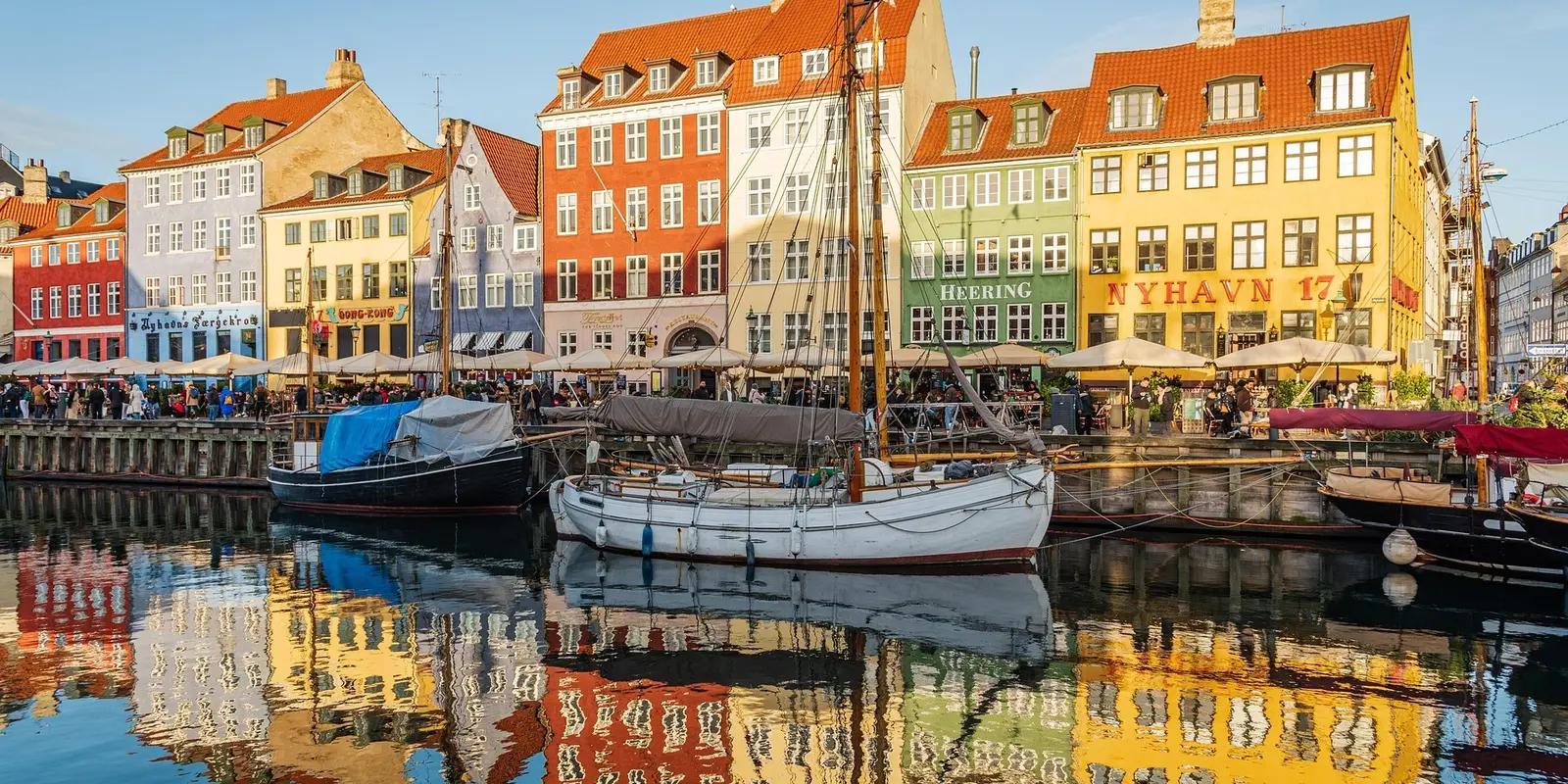 A scenic view of Nyhavn in Copenhagen, Denmark, featuring colorful historic buildings, docked wooden boats, and calm water reflecting the lively harbor.