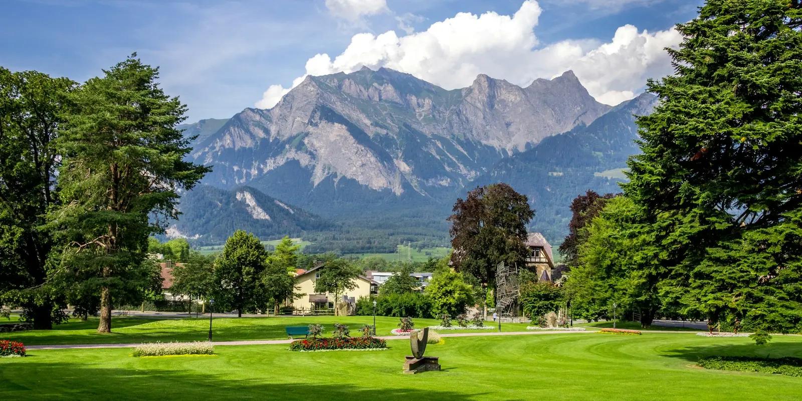 A scenic view of a green park with a mountainous backdrop and a small village nestled at the base of the mountains.