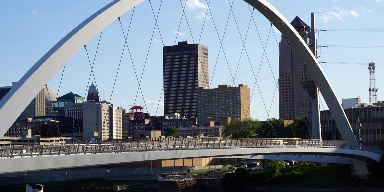 Modern pedestrian bridge with white arch spanning across the river in Des Moines, Iowa, with downtown skyline framed by the structure.