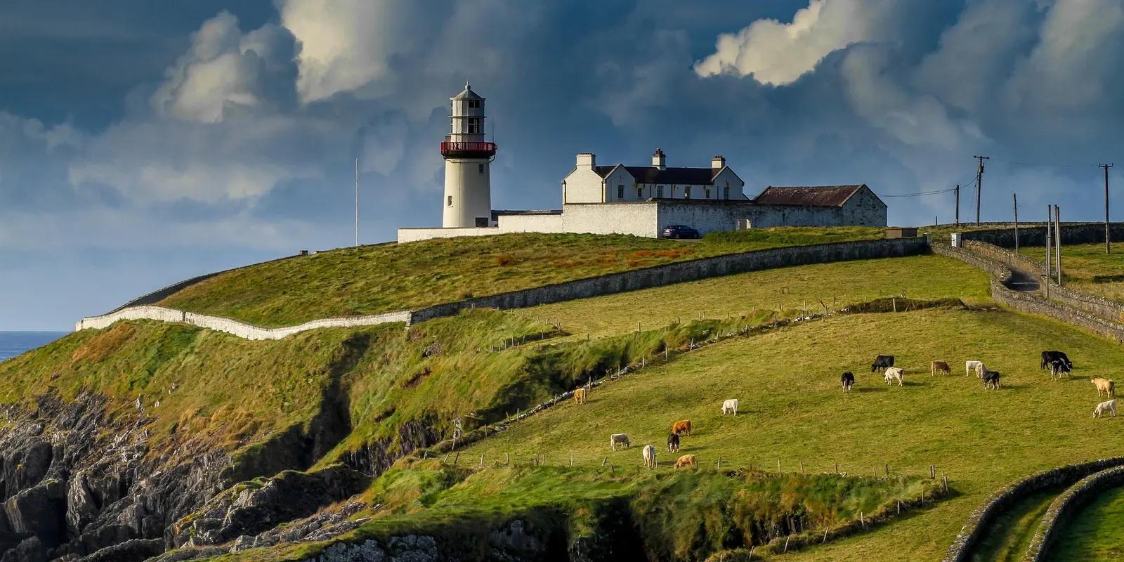 A scenic lighthouse atop a green hillside, overlooking the ocean, with cows grazing under a sky filled with dramatic clouds.