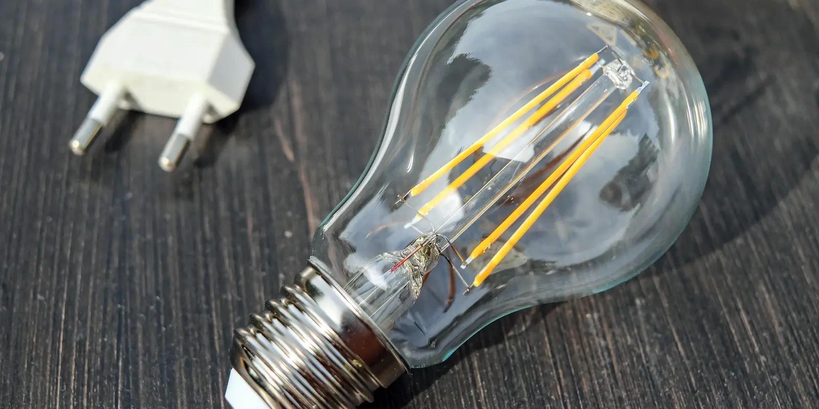A clear LED filament light bulb resting on a dark wooden surface with a two-pronged white plug nearby.