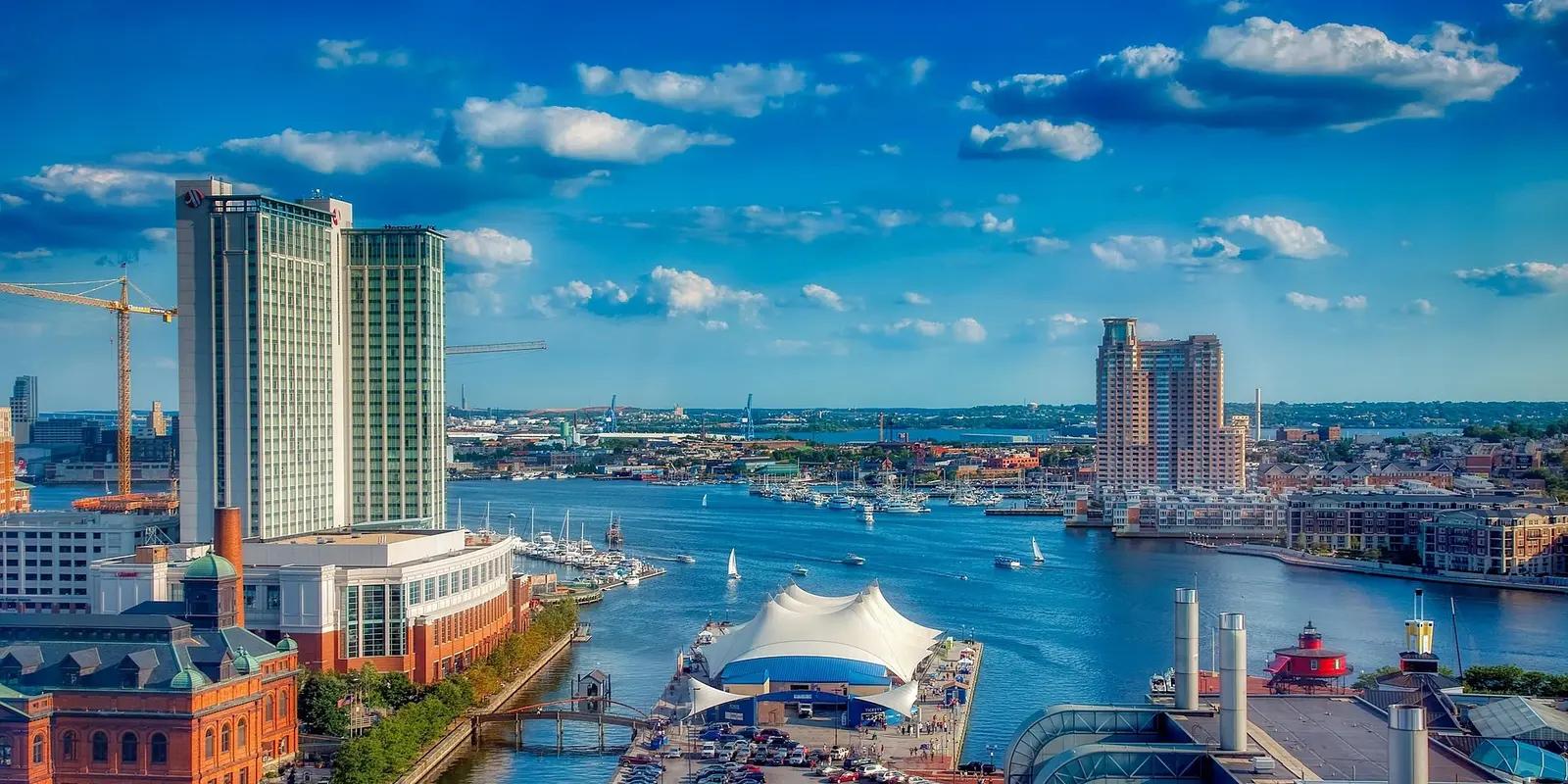Aerial view of Baltimore's Inner Harbor featuring high-rise buildings, waterfront pavilions, and sailboats dotting the blue water under a bright sky.