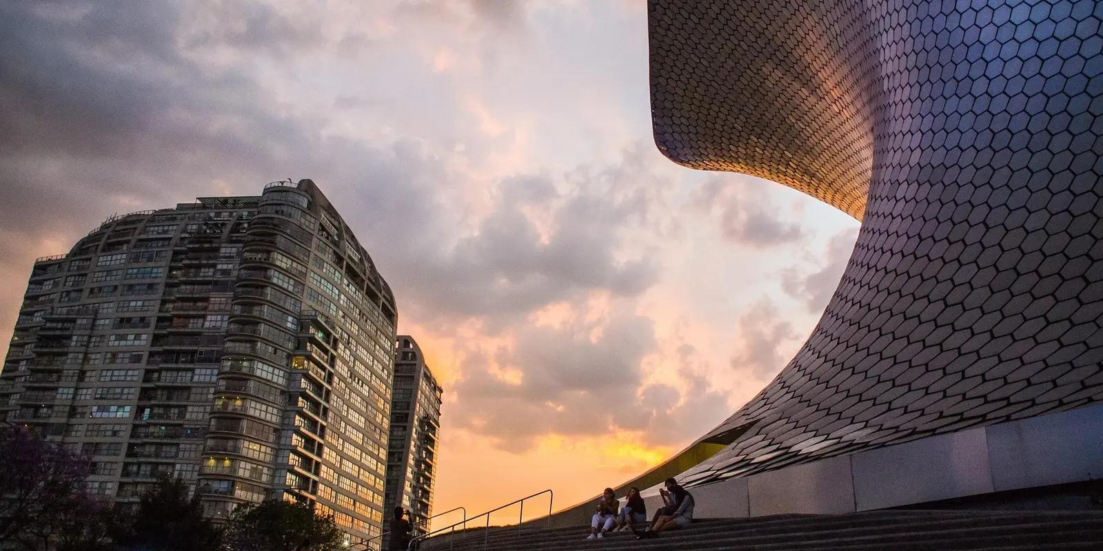 A stunning urban scene at sunset featuring the Soumaya Museum in Mexico City, with its curved, hexagonal-tiled facade reflecting the warm hues of the sky.