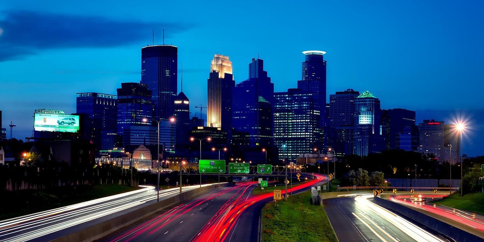 Minneapolis skyline at twilight with illuminated skyscrapers and highway light trails.