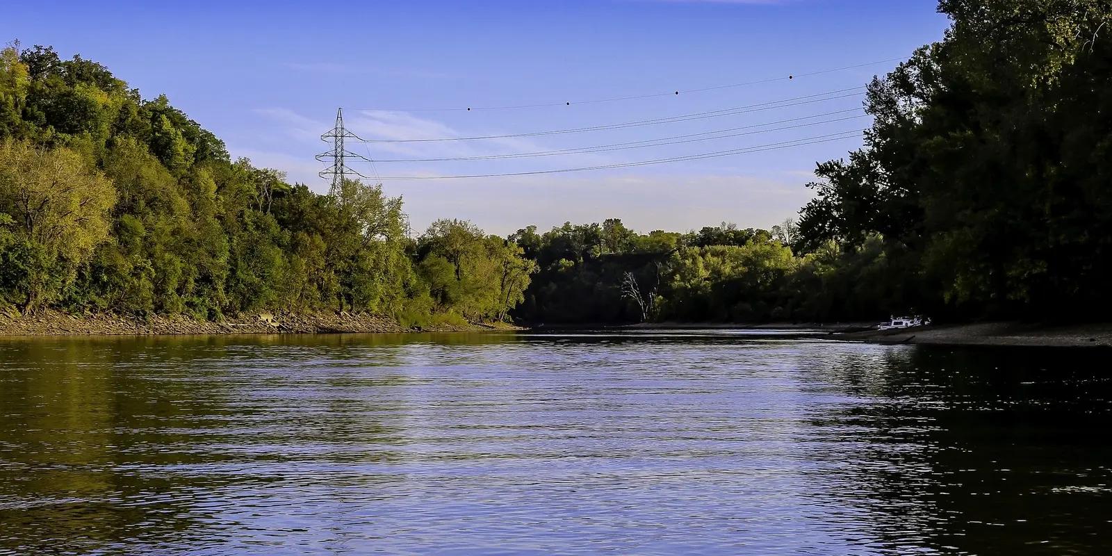 River flowing between tree-lined banks under clear blue sky with distant power lines.