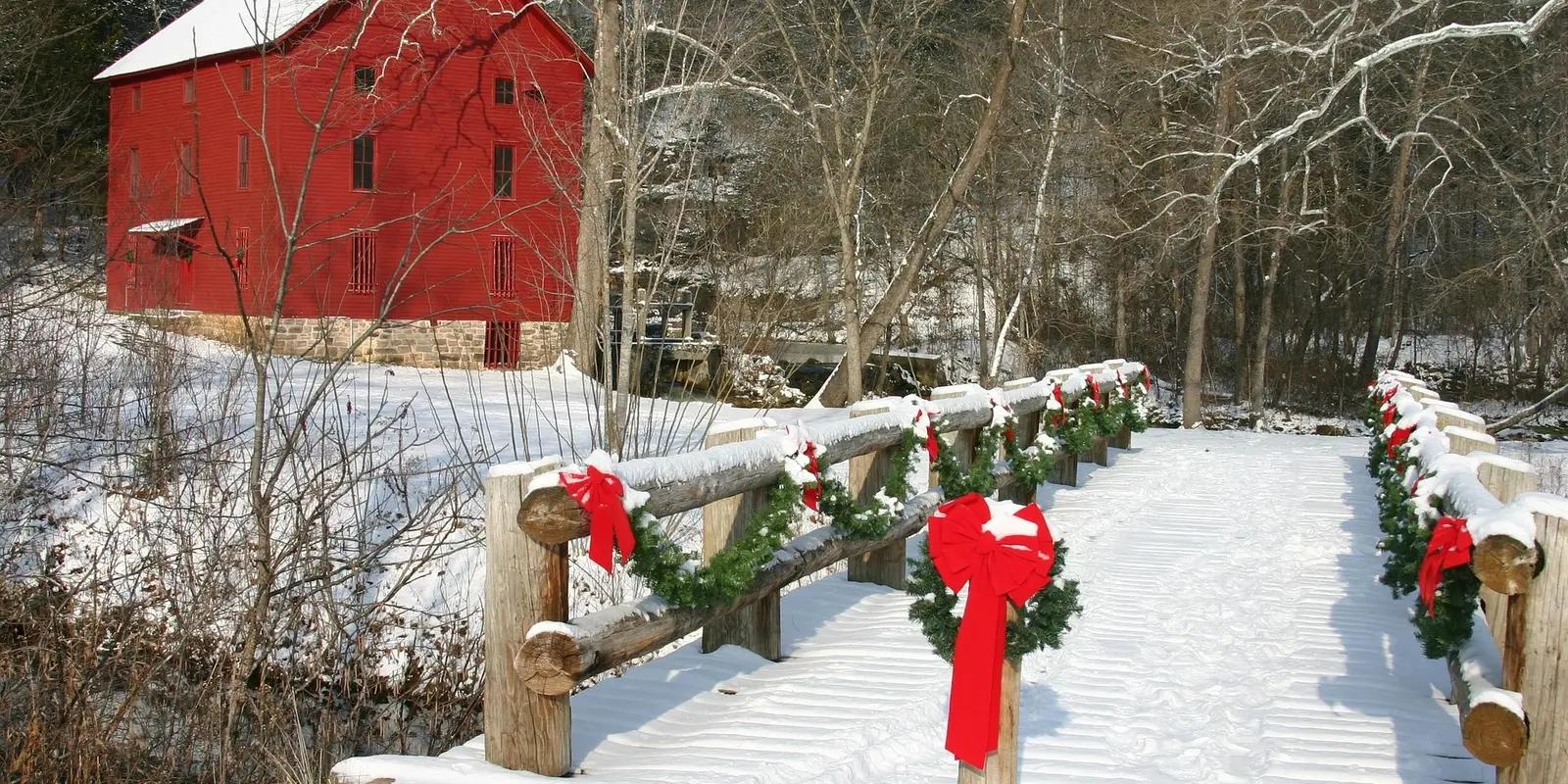 Red barn in winter landscape with snow-covered wooden bridge decorated with Christmas wreaths.