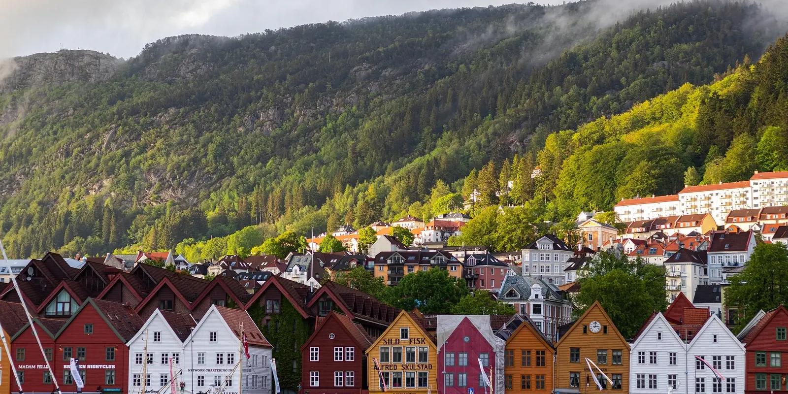 A scenic view of Bryggen in Bergen, Norway, showcasing colorful wooden buildings along the waterfront with a backdrop of lush green mountains and misty clouds.