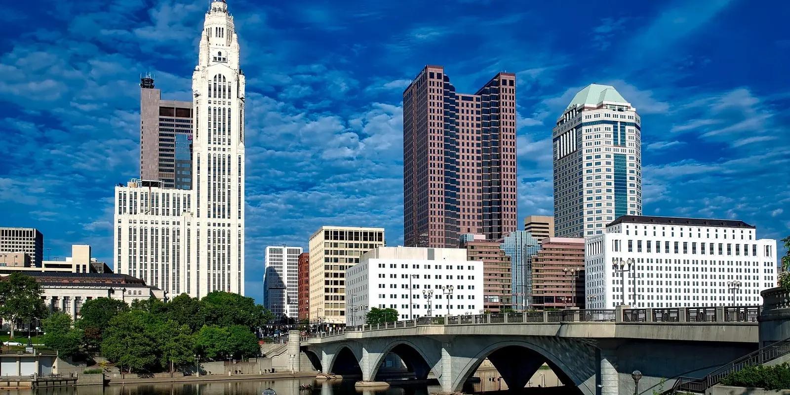 A cityscape of Columbus, Ohio, featuring a mix of modern and historic skyscrapers, a bridge over the Scioto River, and a vibrant blue sky with scattered clouds.