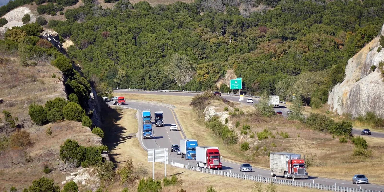 A scenic highway in Oklahoma winding through rolling hills covered in green vegetation, with multiple semi-trucks and cars traveling in both directions under a clear blue sky.