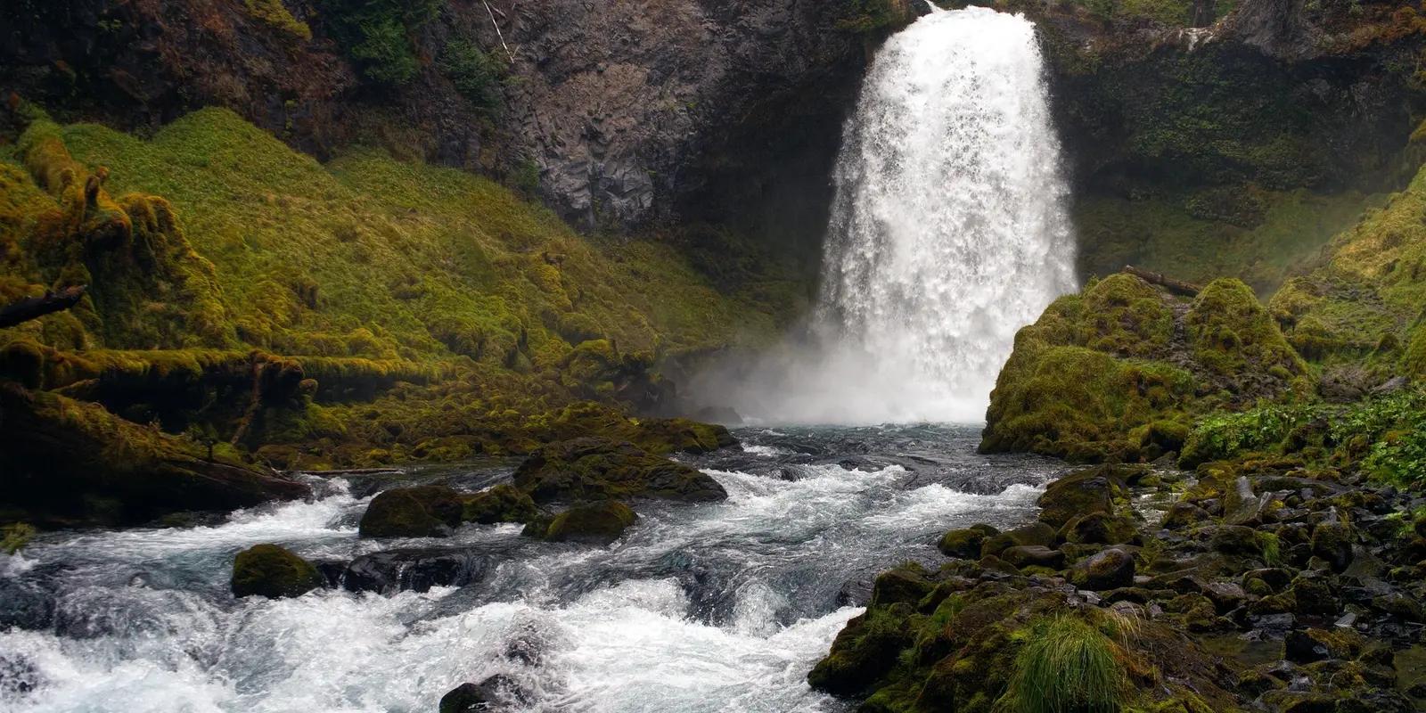 A stunning waterfall in Oregon cascading down a moss-covered rocky cliff into a rushing river, surrounded by lush green vegetation and dense forest.