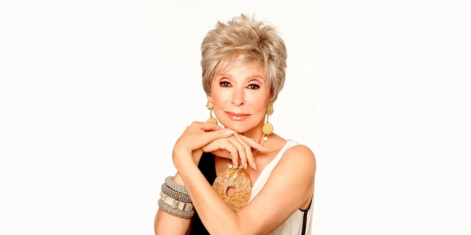 Rita Moreno posing elegantly against white background, wearing gold earrings and statement jewelry, with short silver hair and a white top.