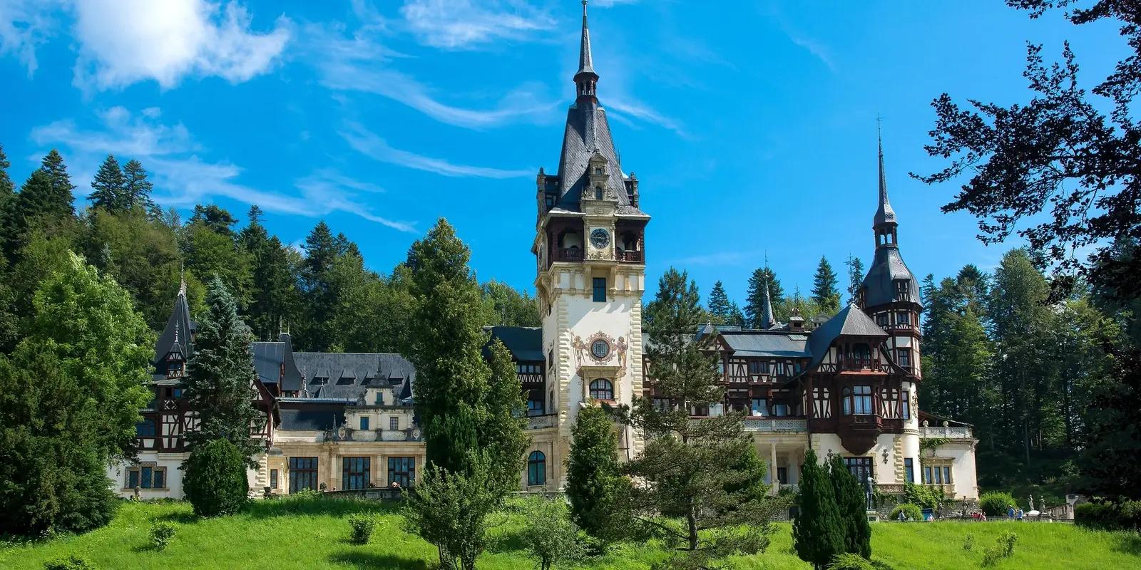 Peleș Castle in Romania with its distinctive Germanic architecture, featuring a tall central clock tower flying the Romanian flag, surrounded by lush green lawns and forested mountains under a bright blue sky.