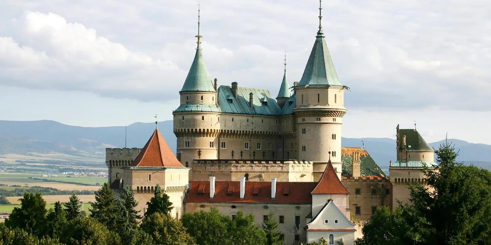 Bojnice Castle with distinctive blue-green spires and turrets set against a backdrop of forests and mountains in the Slovak countryside.
