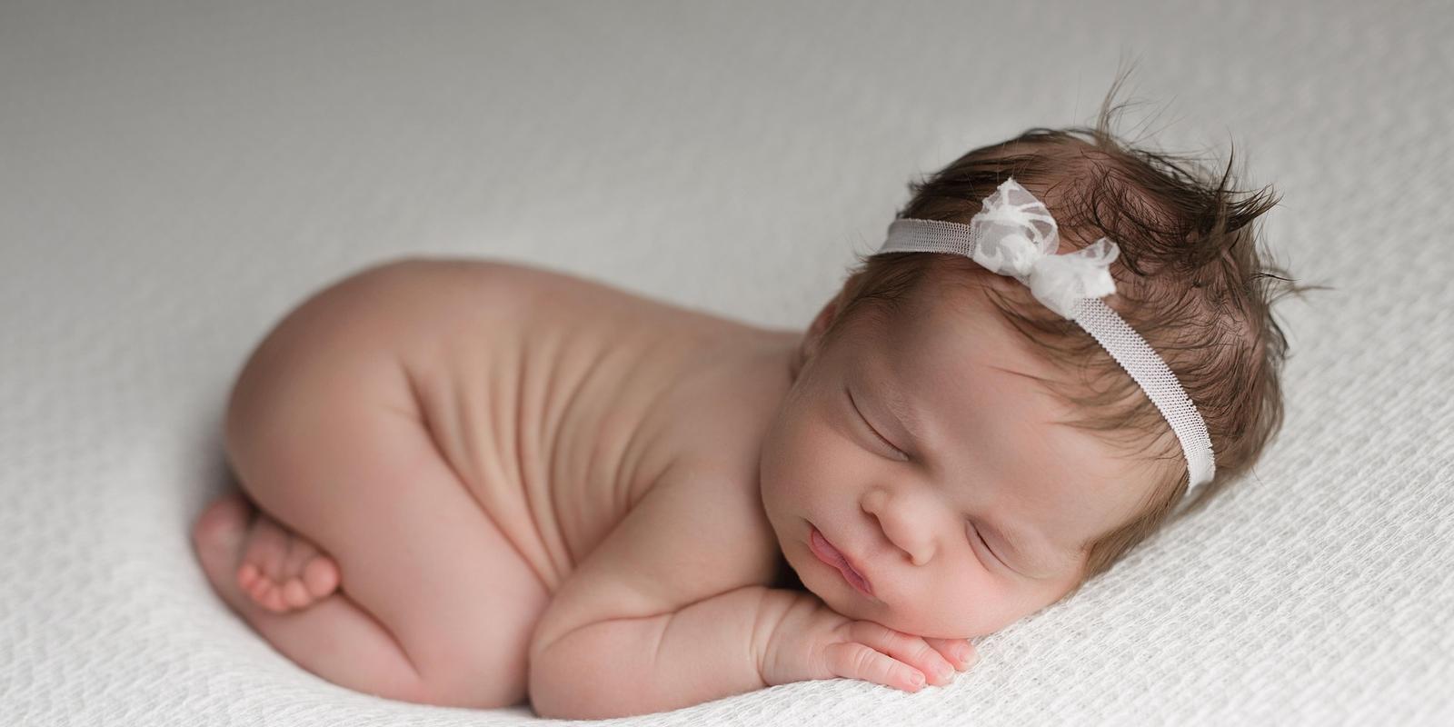 Newborn baby sleeping peacefully on white fabric, wearing a delicate white headband, with soft brown hair visible atop their head.