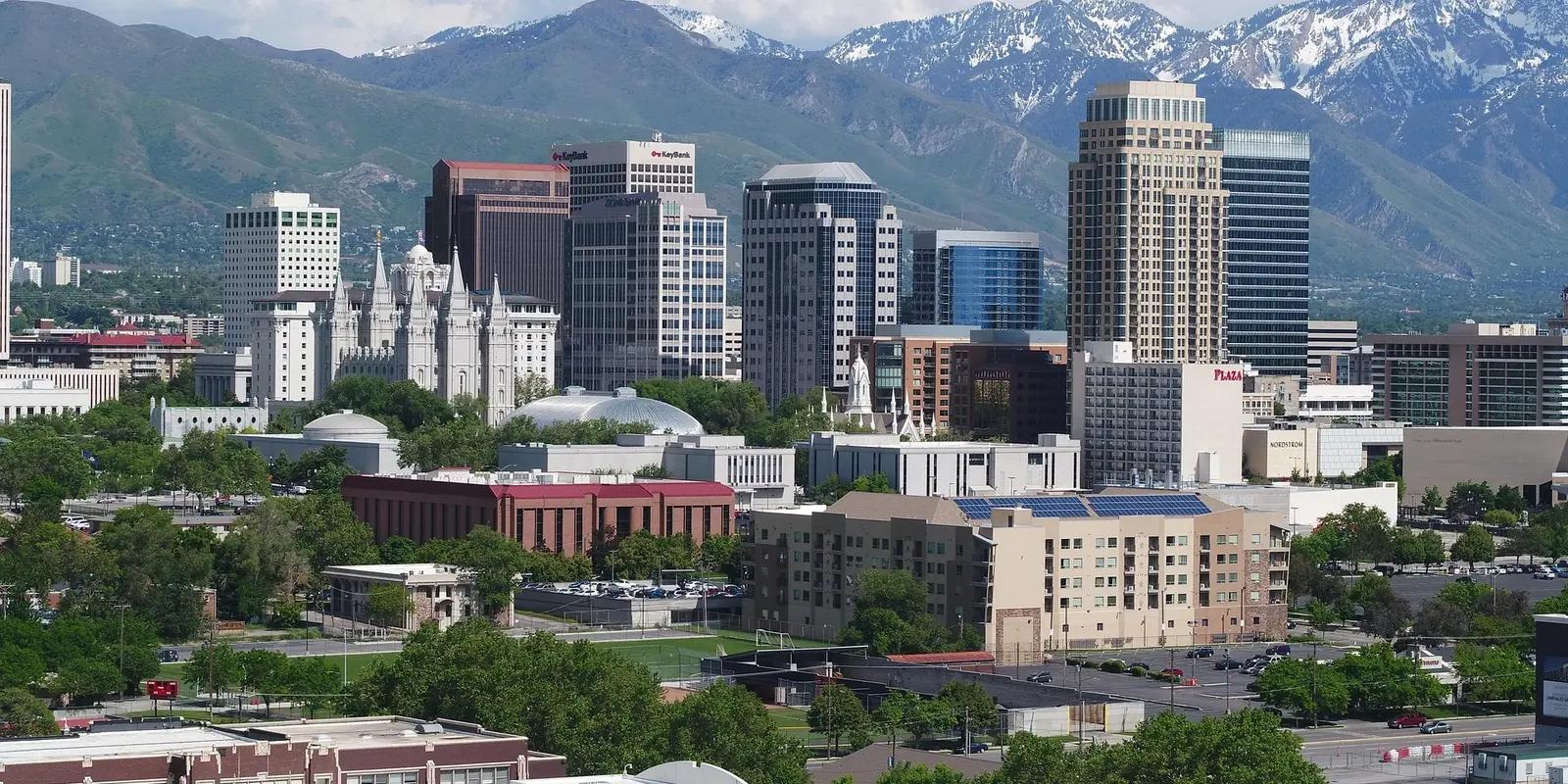 A panoramic view of Salt Lake City, Utah, showcasing the city's skyline with modern skyscrapers, the historic Salt Lake Temple, and the Wasatch Mountains with snow-capped peaks in the background.