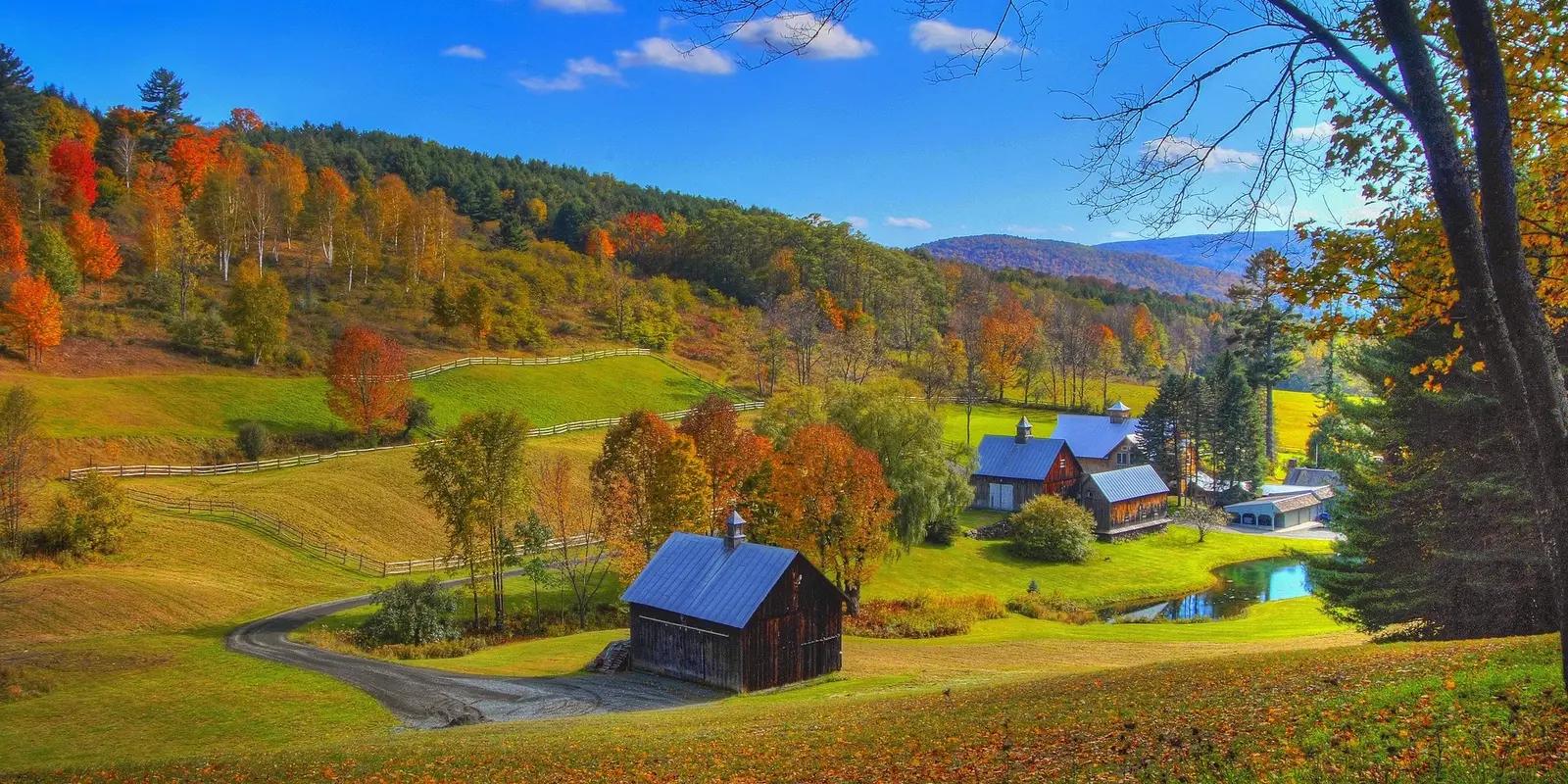 A picturesque autumn landscape in Vermont featuring rolling hills, vibrant fall foliage, a winding country road, and a charming farmhouse with a pond nestled among colorful trees.