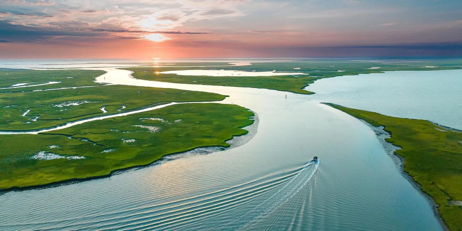 An aerial view of Wachapreague, Virginia, at sunset, showcasing winding waterways weaving through lush green marshlands, with a small boat leaving ripples in the calm coastal waters.