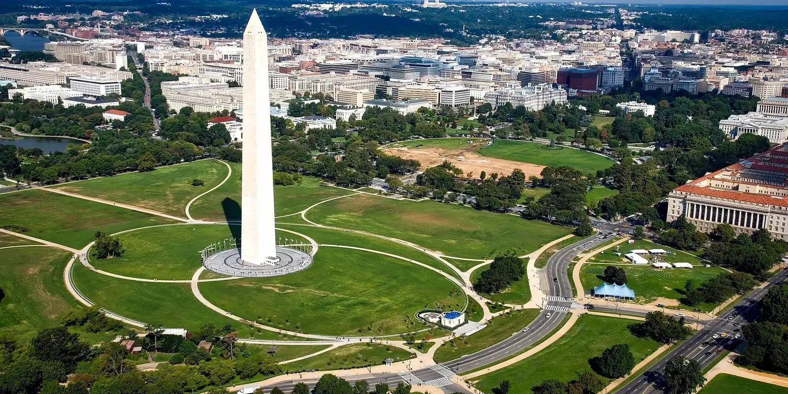 An aerial view of the Washington Monument in Washington, D.C., surrounded by the National Mall's green parkland, intersecting pathways, and city buildings in the background.