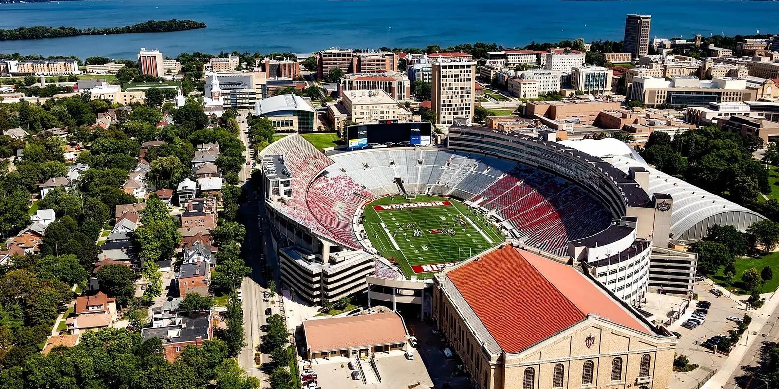 An aerial view of Madison, Wisconsin, showcasing Camp Randall Stadium with its red and white seating, surrounded by city buildings, lush greenery, and a scenic lake in the background.