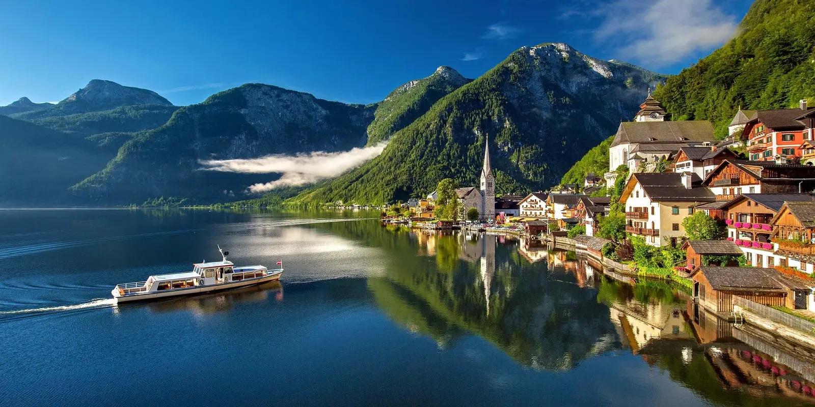 Hallstatt, Austria, featuring a scenic lakeside village with traditional houses, a church steeple, and mountains in the background.