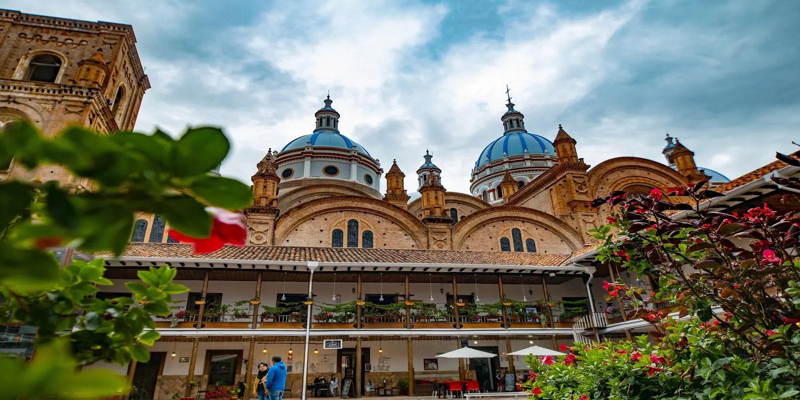 The blue domes of the Cuenca Cathedral in Cuenca, Ecuador, viewed from a courtyard with plants and people.
