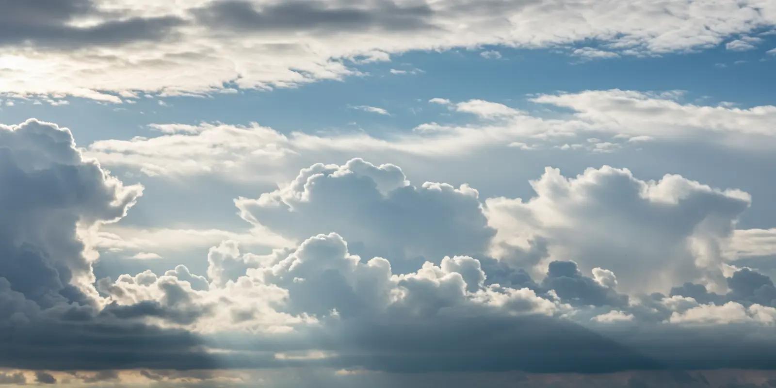 A dynamic sky filled with various types of clouds. Large, puffy white cumulus clouds with darker bases contrast against patches of clear blue sky. Wispy cirrus clouds are visible higher up, and some areas show darker, more diffuse clouds.