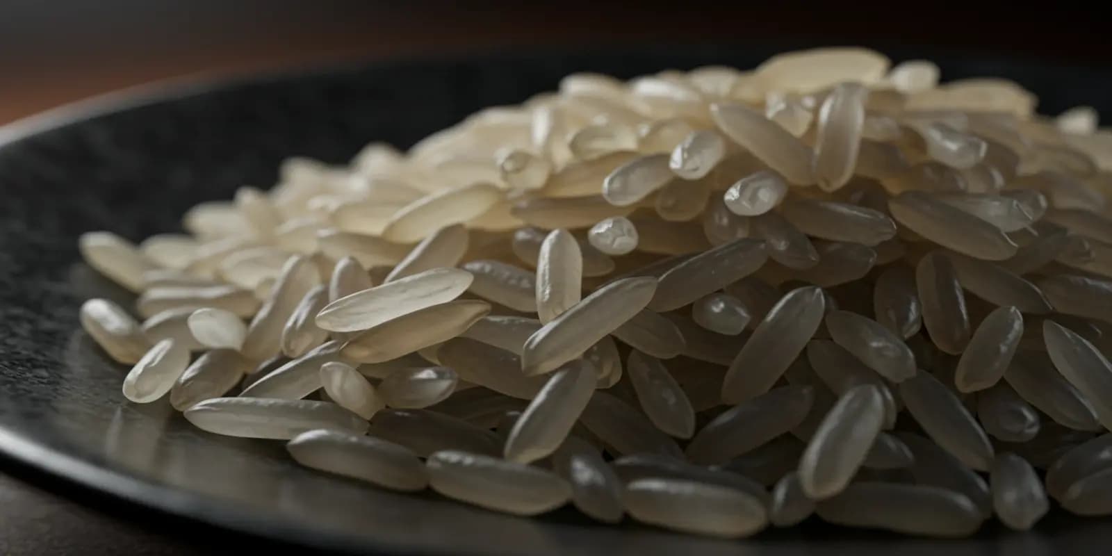 A close-up of a pile of long, slender grains of parboiled rice on a dark, slightly textured surface. The rice grains are translucent and have a light golden hue.