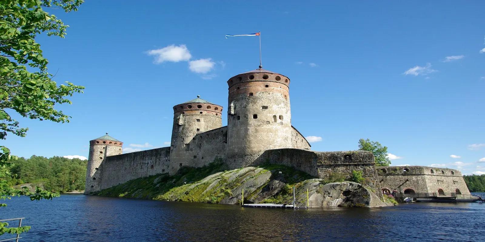 Olavinlinna Castle in Savonlinna, Finland, featuring a medieval stone fortress with towers and a flag, situated on a lake with a clear blue sky.