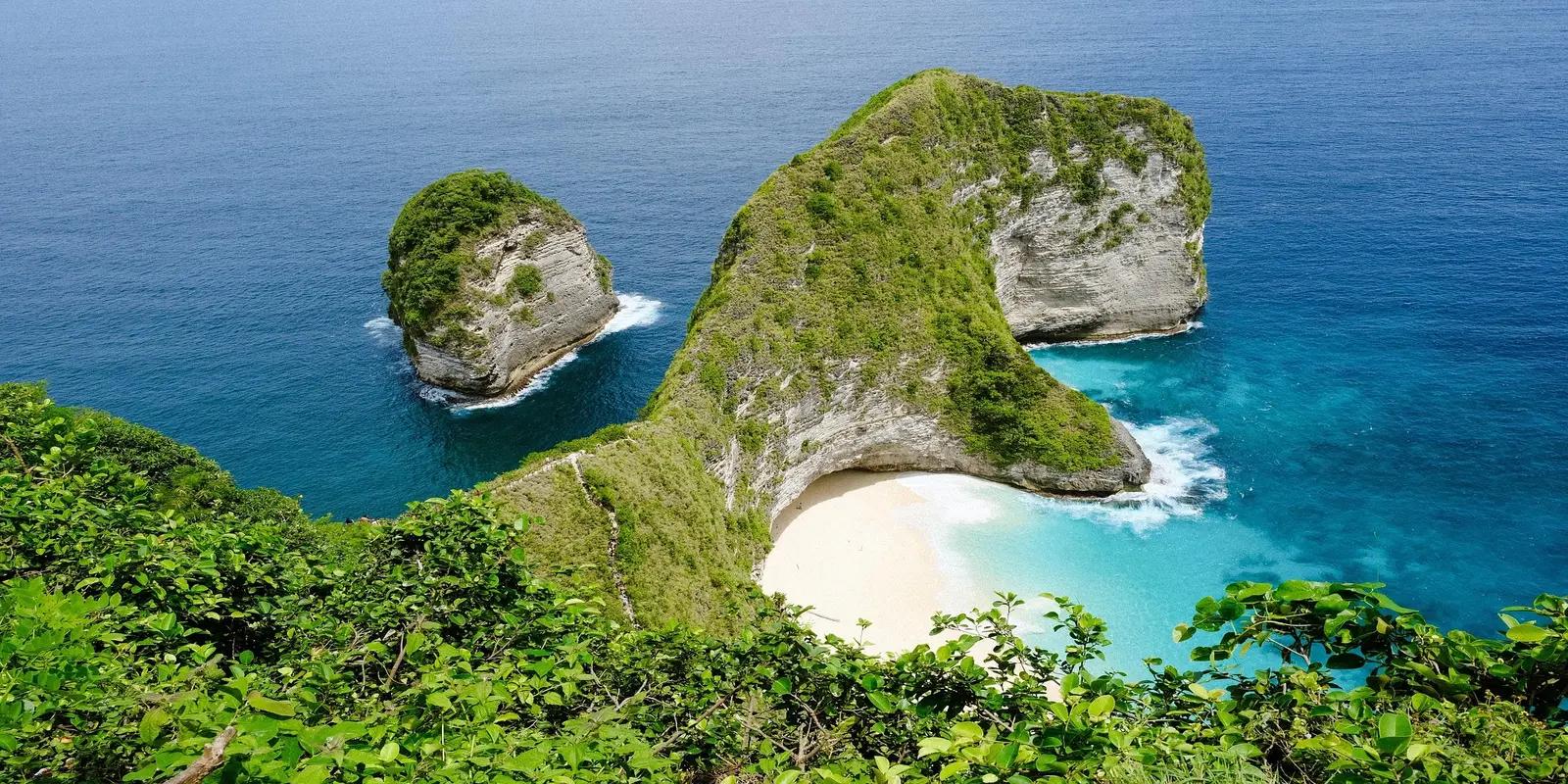 Aerial view of Kelingking Beach on Nusa Penida Island, Bali, featuring a T-Rex shaped cliff covered in greenery, a white sandy beach, and turquoise blue water.
