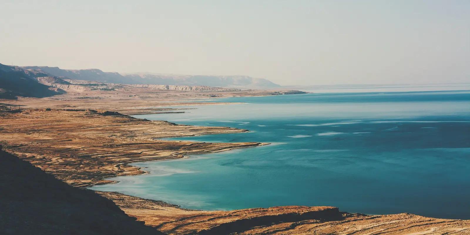 Scenic view of the Dead Sea, featuring a shoreline with layered, dry terrain meeting the turquoise water.
