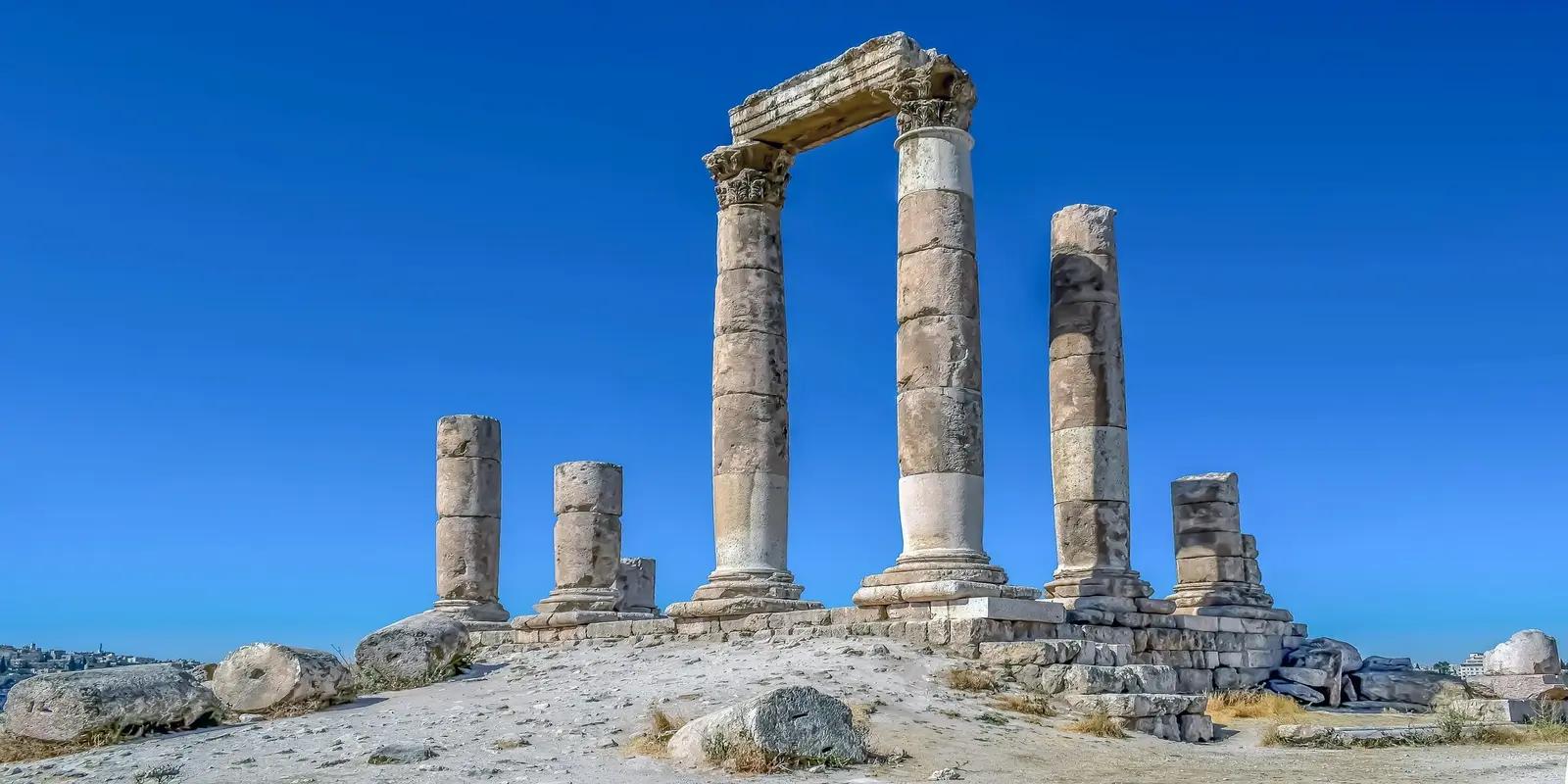 The ruins of the Temple of Hercules at the Amman Citadel in Amman, Jordan, featuring large stone columns and a partially reconstructed arch against a clear blue sky.