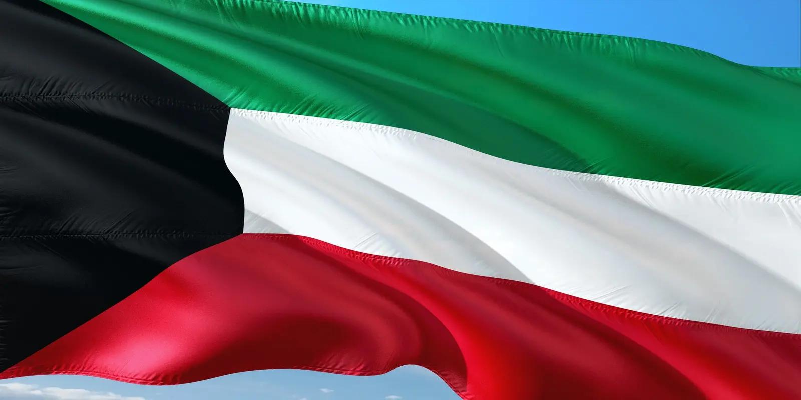 The flag of Kuwait, featuring horizontal stripes of green, white, and red, with a black trapezoid on the hoist side. The flag is shown waving against a blue sky with faint clouds.