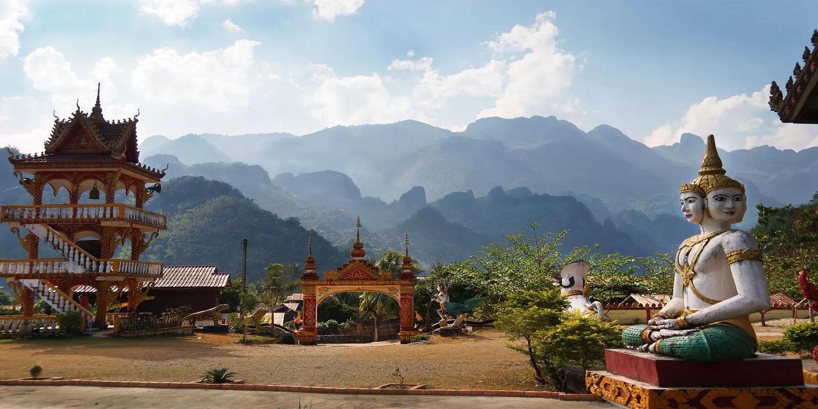 A Buddhist temple in Laos featuring a golden stupa, ornate gates, and a statue of a multi-faced Buddha figure.
