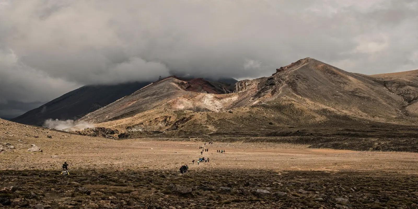 Hikers on a trail through a dramatic volcanic landscape in Tongariro National Park, New Zealand. Barren, multi-colored mountains with a central crater are partially obscured by heavy, low-hanging clouds.