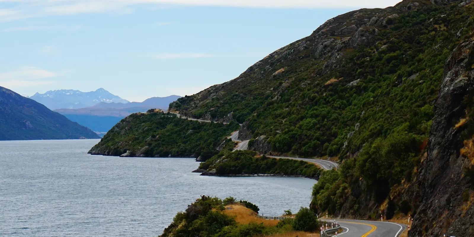 Scenic coastal road winding along a lush green hillside beside a lake in New Zealand, with mountains in the background.