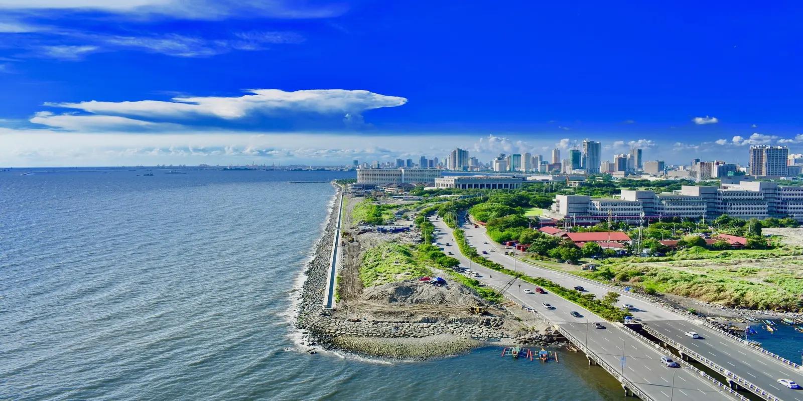 Aerial view of Manila, Philippines, featuring the coastline along Manila Bay with a road running parallel, leading to the city skyline in the distance.